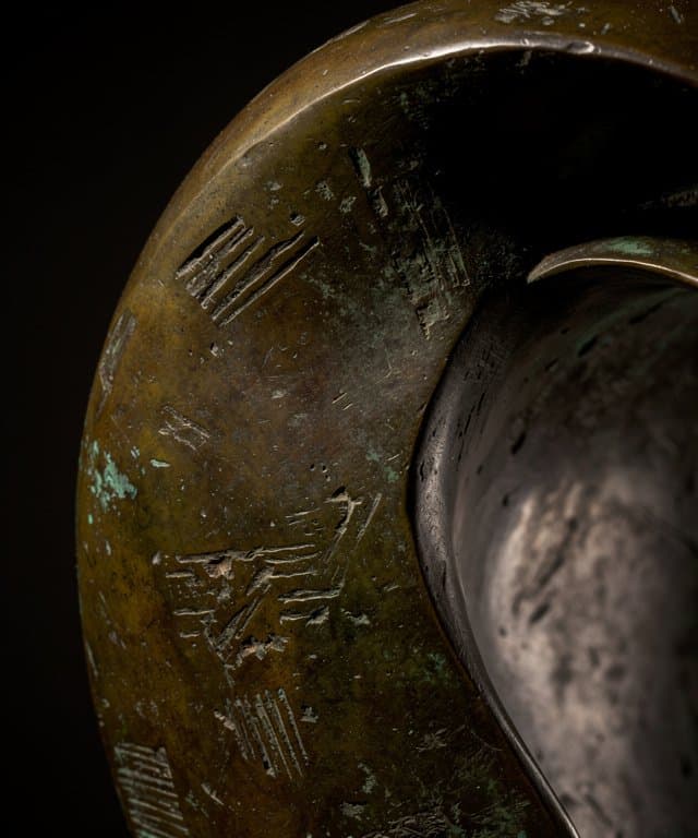 Macro fine art photograph of a weathered bronze sculpture, tightly cropped abstract curved sculptural surface with visible hand-worked irregular patina in dark umber, olive and subdued verdigris, raking museum light revealing tactile texture, deep quiet background, no people, no text