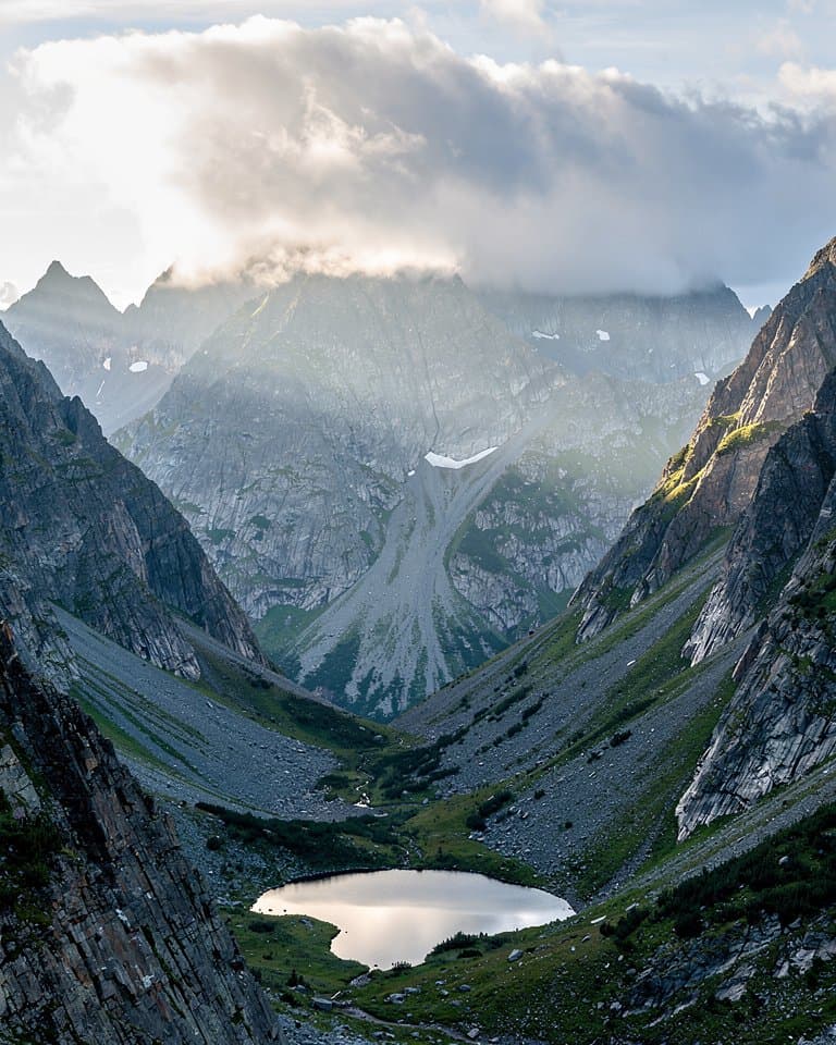 Vertical large-format landscape photograph looking through a high North Cascades mountain pass, layered slate peaks and a small alpine tarn below, rain cloud lifting into cool early morning light, delicate mist on the slopes, believable natural color and finely resolved rock texture, no people, no structures, no text. Portrait frame.
