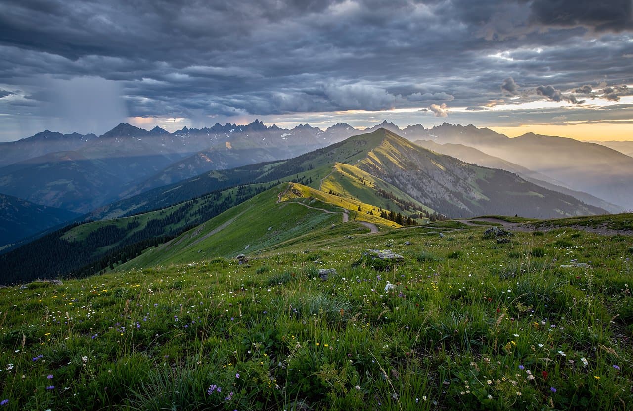 Wide landscape photograph of a long Wallowa Mountains ridge in eastern Oregon just after rain, deep green alpine meadow in foreground, broken cloud shadow crossing distant blue-grey peaks, a narrow glimpse of low evening sunlight, lifelike weather, large-format fine detail, natural colors, no people or structures, no text.