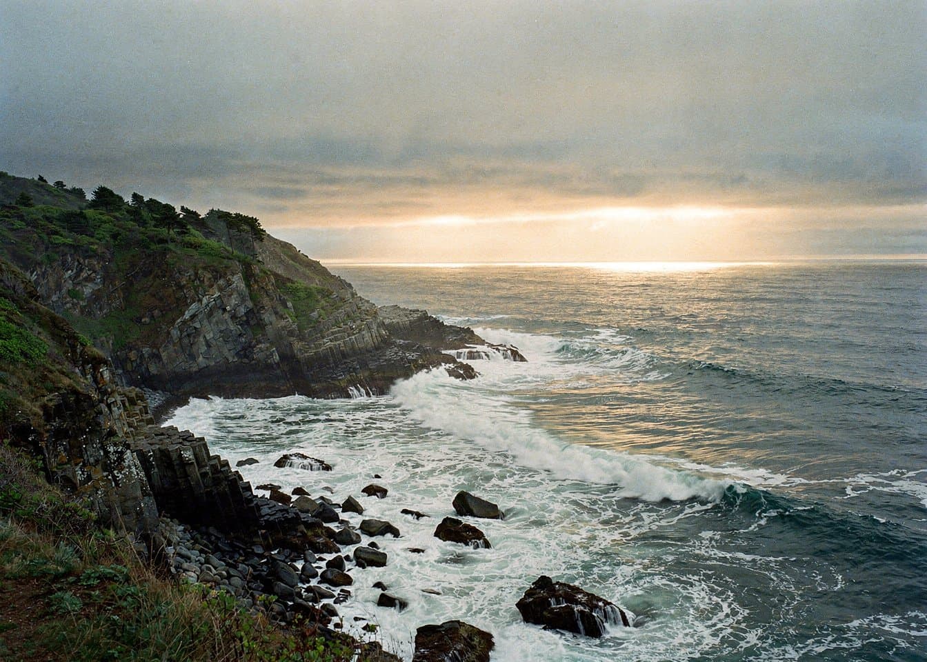 Large-format landscape photograph of a rugged Oregon Pacific headland at late afternoon, dark basalt ledges, white surf threading into a quiet cove, low marine cloud with one band of clear warm light on the sea, natural subdued colors, fine film grain and exceptional detail, no people, no buildings, no text. Wide horizontal frame.