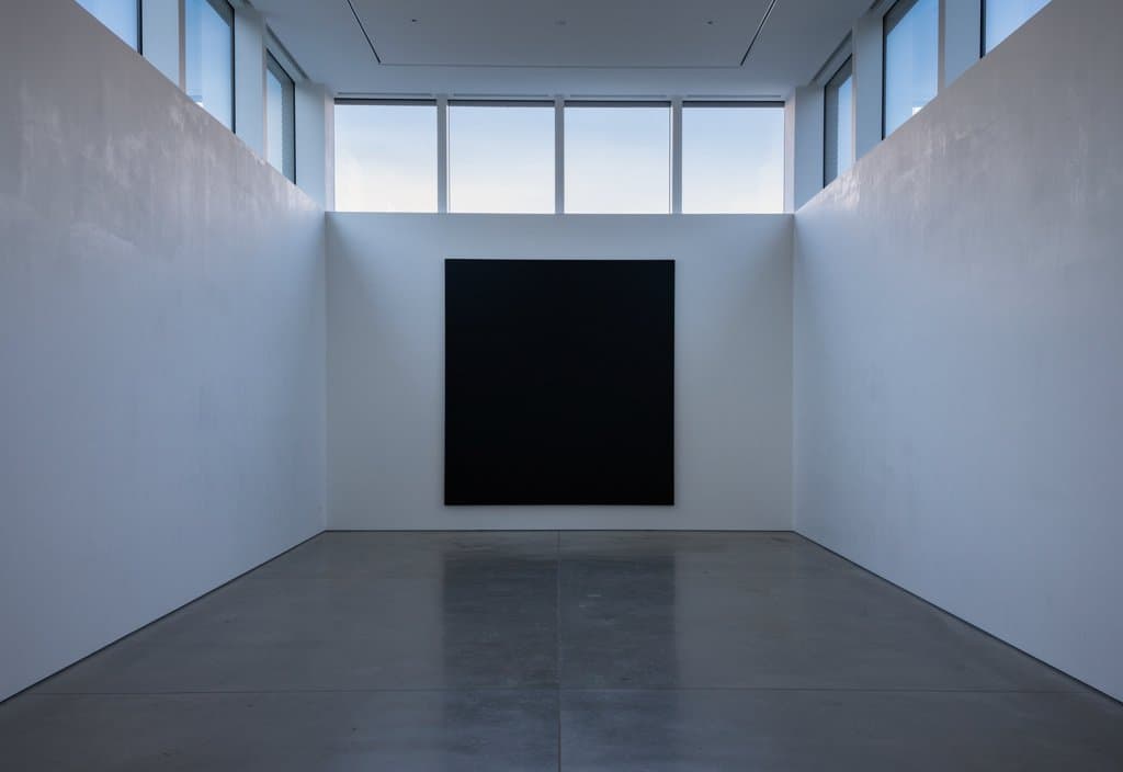 Quiet contemporary art photography, wide interior view of a completely empty bare white exhibition hall at dusk, a single very large nearly black matte canvas on the far wall, soft fading blue evening light through high windows, pale plaster walls, polished concrete floor, spare architectural composition, realistic fine art installation photograph, no people, no text, no signage
